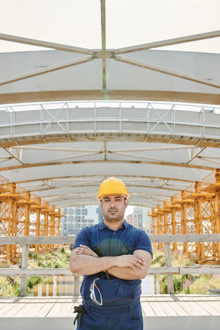 Male construction worker with crossed arms at a bridge site with orange scaffolding.